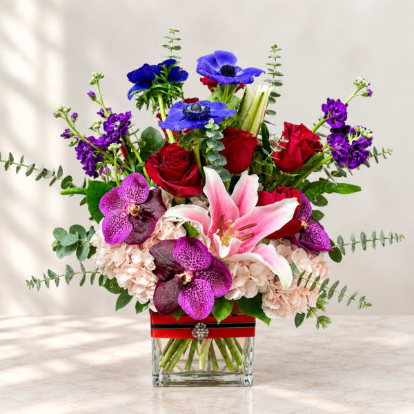 Mixed bouquet of red roses, pink lilies, purple orchids, and blue anemones in a glass vase