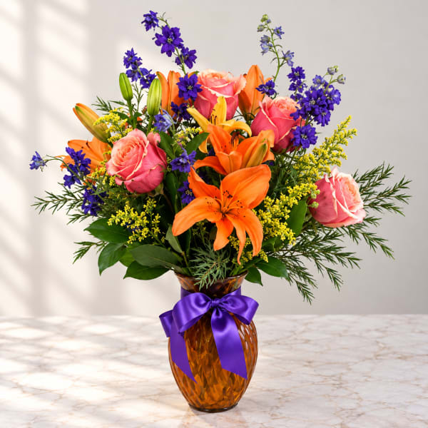 Bouquet of orange lilies, pink roses, and purple flowers in an amber vase with a purple ribbon
