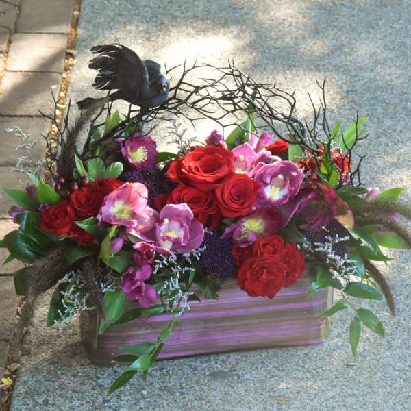 Low rectangular arrangement of red roses and purple flowers with a black bird on twig accents