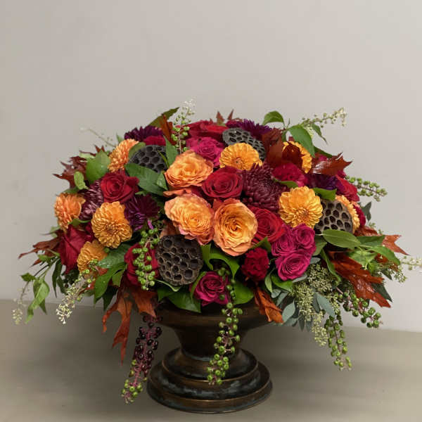Low arrangement of orange and red roses with dark accent flowers in a bronze pedestal bowl