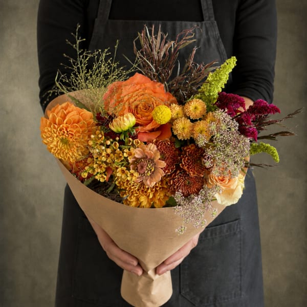 Handheld bouquet of orange, yellow, and burgundy flowers wrapped in kraft paper