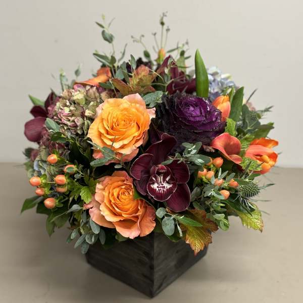 Low arrangement of orange roses, dark burgundy orchids, and mixed blooms in a square dark wooden vase