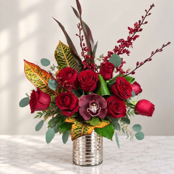 Red roses arranged in a silver vase with burgundy orchids and patterned leaves