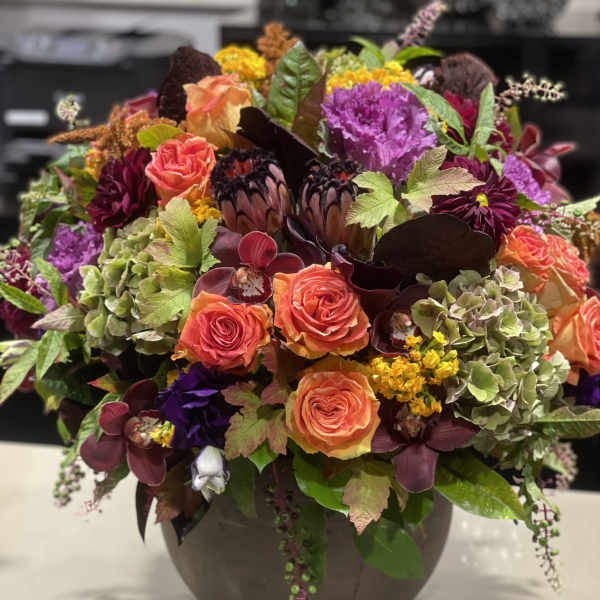 Lush autumn-toned arrangement with orange roses, protea, orchids and mixed blooms in a round ceramic vase