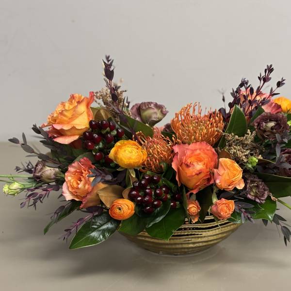 Low centerpiece of orange roses, pincushion protea, and dark berries in a striped gold bowl