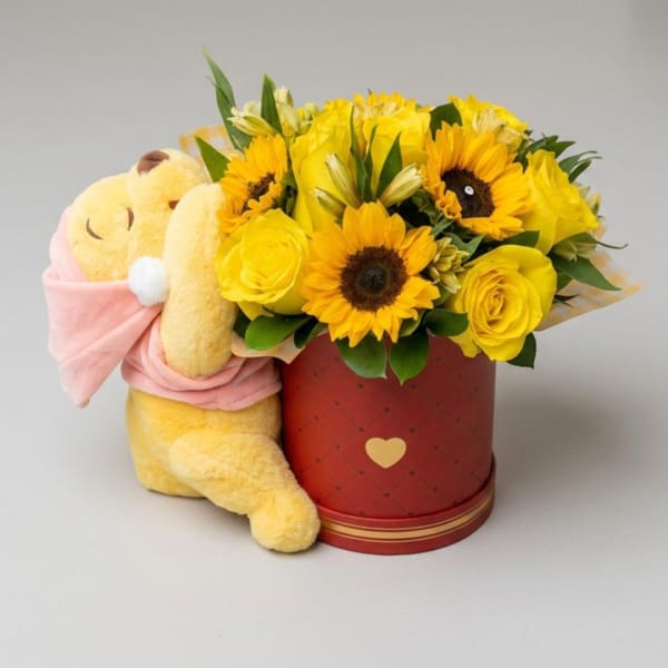 Yellow flower arrangement in a red box beside a plush teddy bear