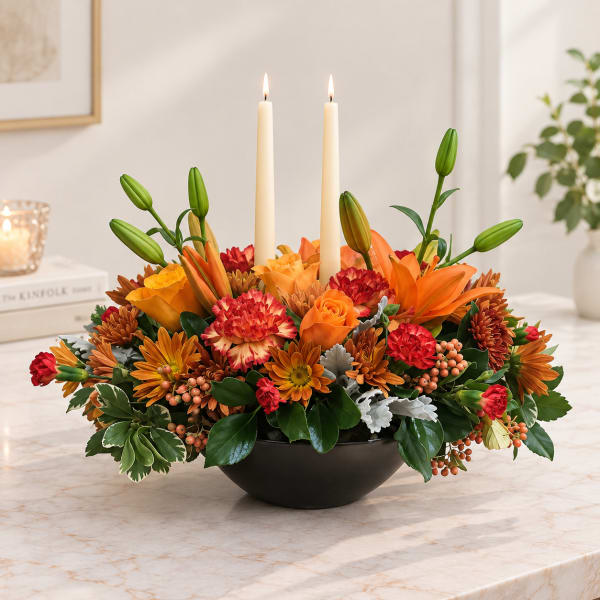Orange and red floral centerpiece with two lit taper candles in a black bowl
