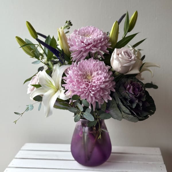 Pink chrysanthemums and lilies arranged with roses in a purple glass vase.