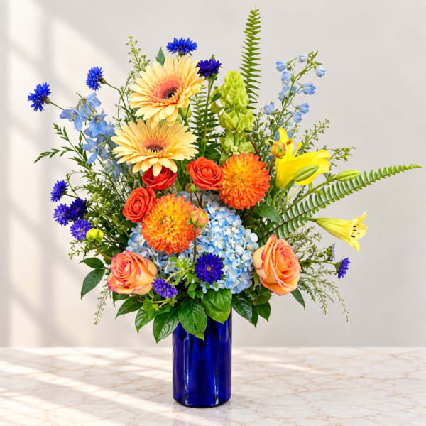 Colorful mixed bouquet in a blue glass vase with roses, gerberas, lilies, and blue flowers