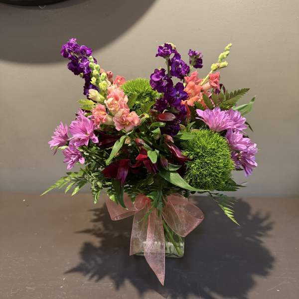 Mixed bouquet of purple, pink, and green flowers in a glass vase with a pink ribbon