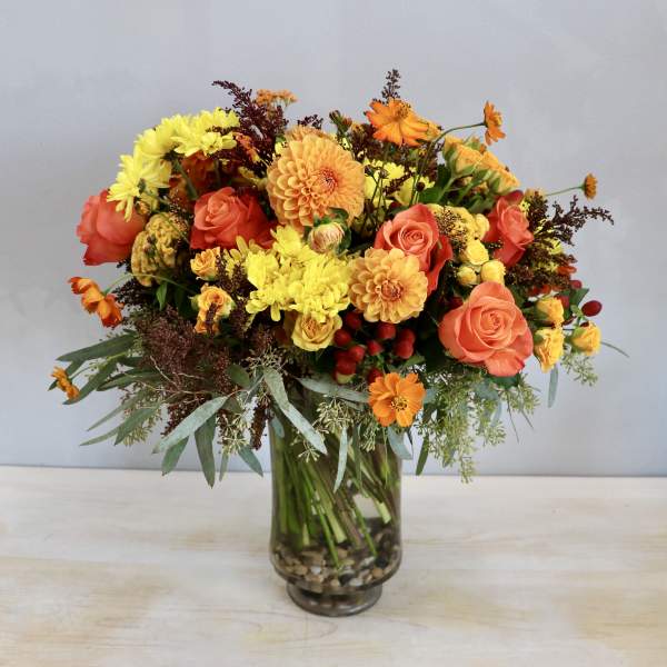 Tall mixed bouquet of orange roses, yellow mums, and dahlias in a clear glass vase with stones.