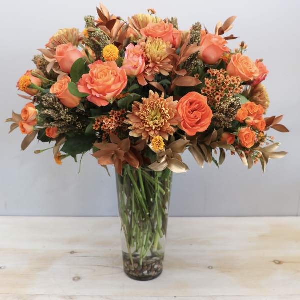Tall vase arrangement of orange roses and chrysanthemums with bronze accents in a clear glass cylinder