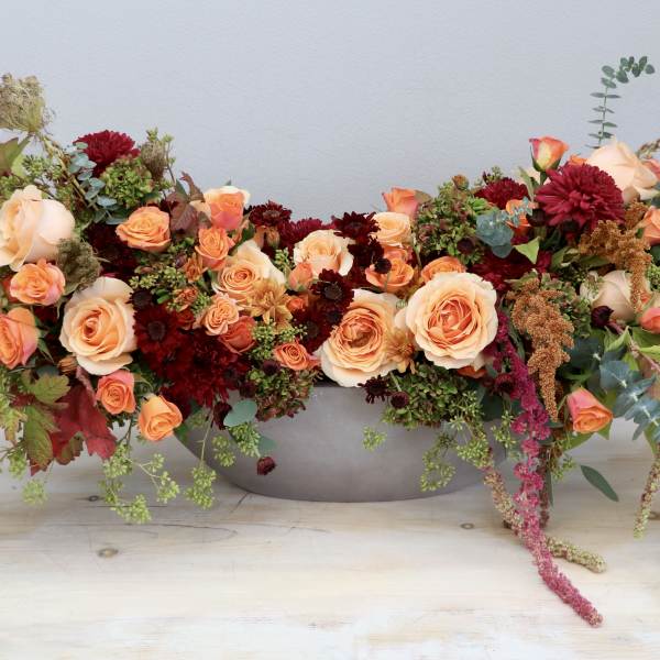 Low oval centerpiece of peach roses and burgundy flowers in a gray bowl-shaped container