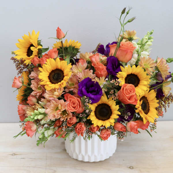 Bright arrangement of sunflowers, orange roses, and purple blooms in a white ceramic vase