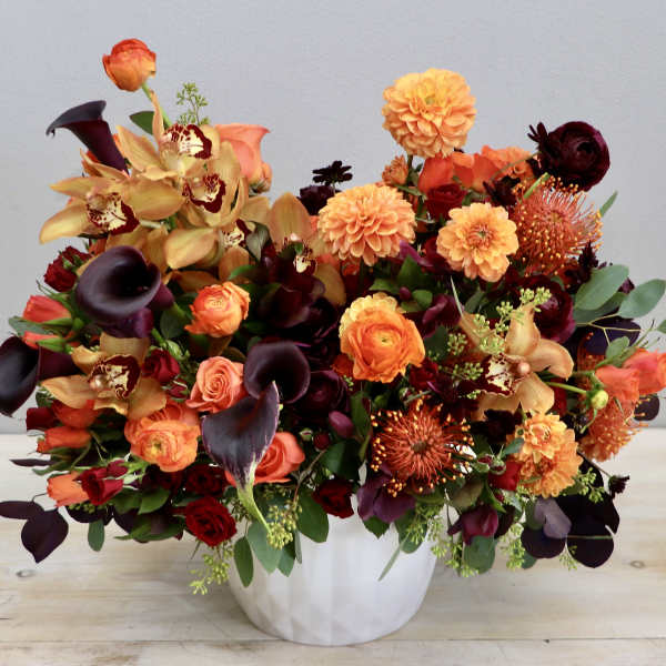 Low white vase filled with orange and burgundy flowers including roses, orchids, dahlias, and calla lilies
