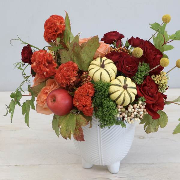 Autumn arrangement with red and orange flowers, mini pumpkins, and an apple in a white ceramic vase