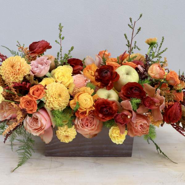 Low rectangular arrangement of orange, yellow, and red flowers with apples in a metal container