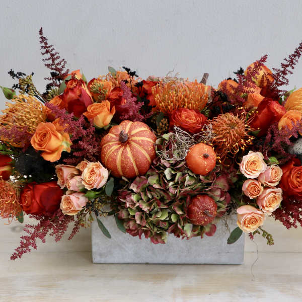 Low rectangular centerpiece with orange and red roses, protea, hydrangea, and small pumpkins.