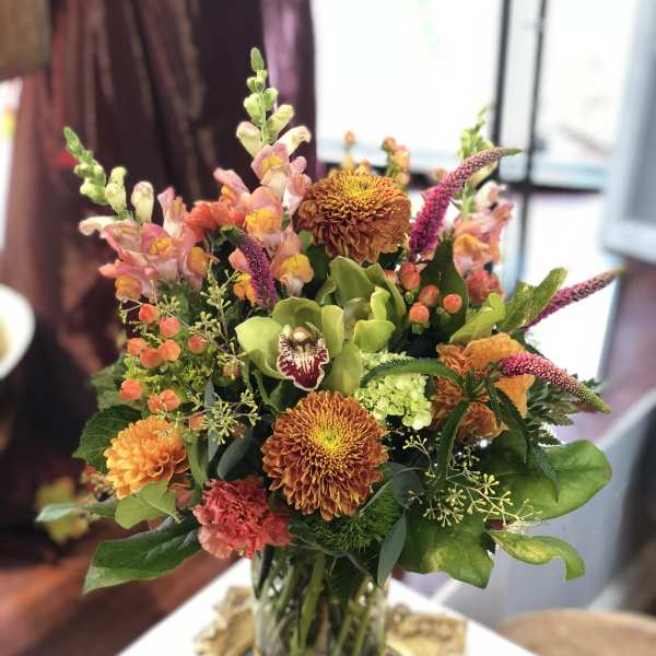 Tall mixed arrangement with orange mums, snapdragons, and green orchid in a glass vase with gold band