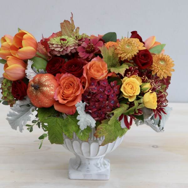 Low orange and red flower arrangement with roses and tulips in a white pedestal bowl.