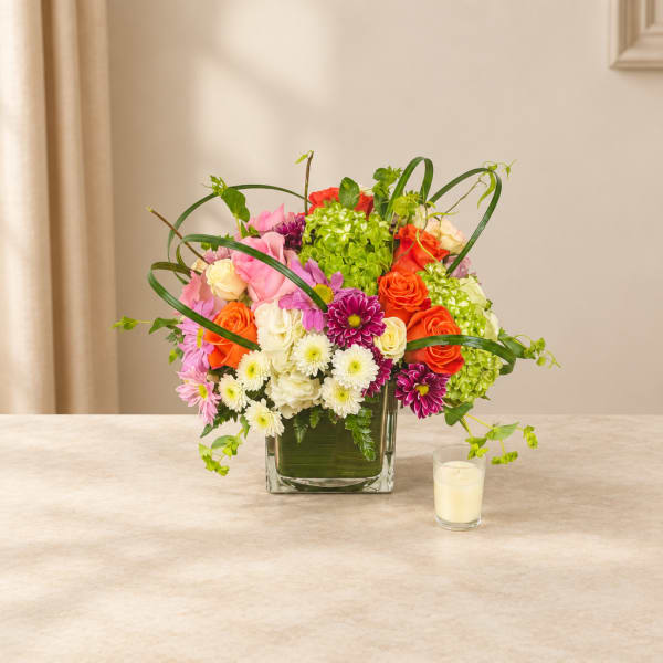 Colorful mixed flower arrangement in a square glass vase with a candle beside it