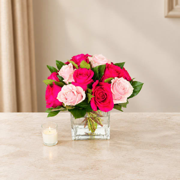 Pink and pale rose bouquet in a clear square vase with a small candle nearby