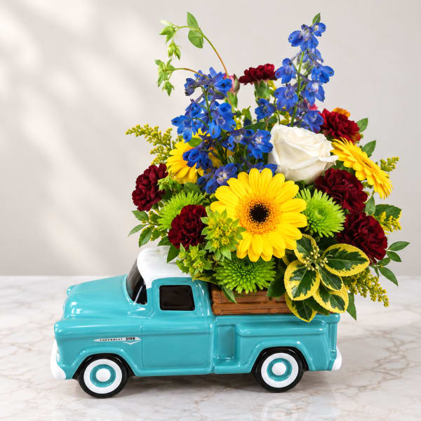 Mixed bouquet in a turquoise truck-shaped container with yellow, blue, red, and white flowers