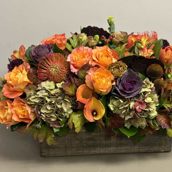 Low centerpiece of orange roses and mixed autumn flowers in a rustic wooden box.