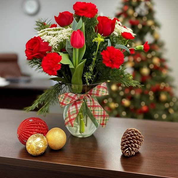 Red carnations and tulips in a glass vase with plaid bow, with ornaments and a pinecone on the table.