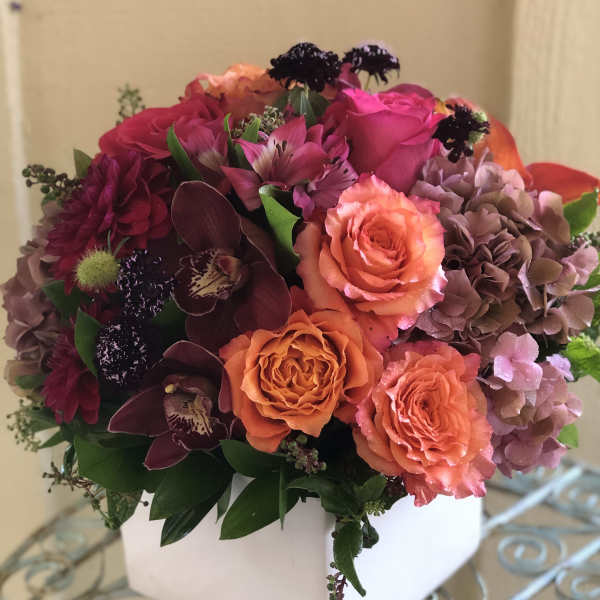 Low cube arrangement of orange roses, pink blooms, and burgundy orchids in a white square vase