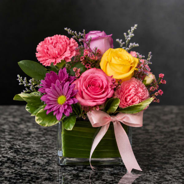 Mixed bouquet of roses, carnations, and purple daisies in a glass vase with a pink ribbon