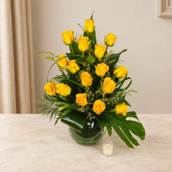 Yellow roses arranged in a glass vase with tropical greenery