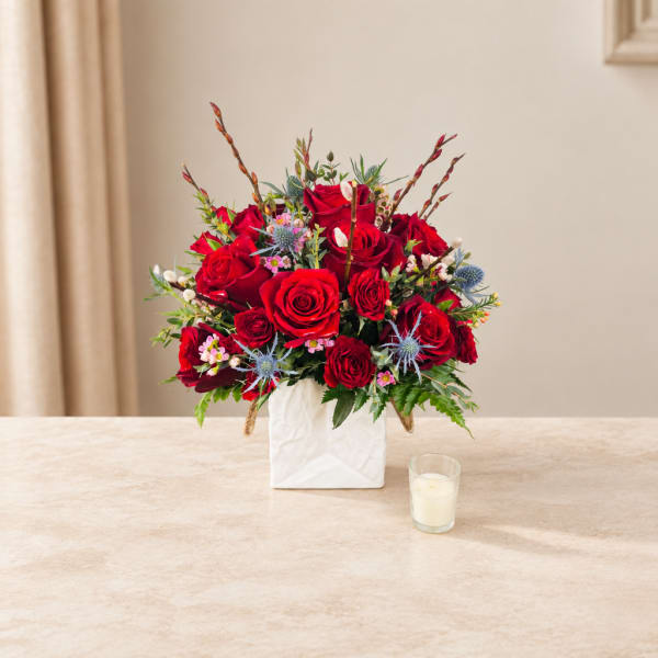 Red rose arrangement in a white square vase with a small candle nearby