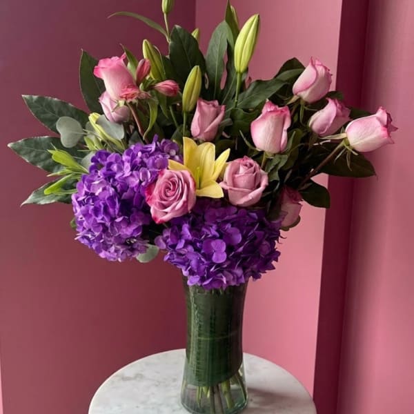 Pink roses, purple hydrangeas, and yellow lilies in a tall glass vase