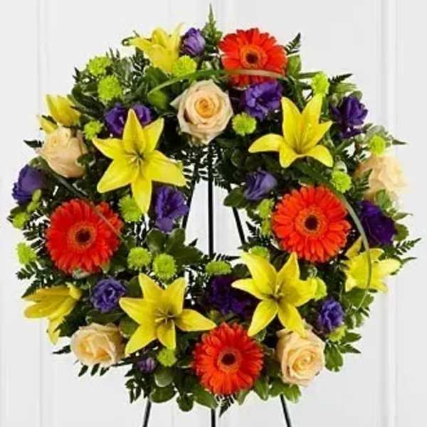 Colorful standing wreath with yellow lilies, orange gerbera daisies, roses, and purple blooms on an easel.