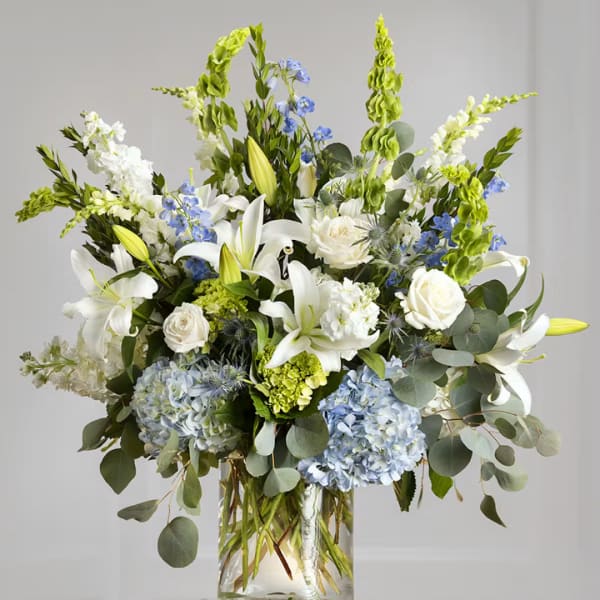 Tall bouquet of white lilies, roses, and blue hydrangeas in a glass vase