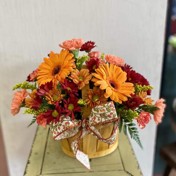 Orange and red mixed flowers arranged in a wooden basket with a patterned bow