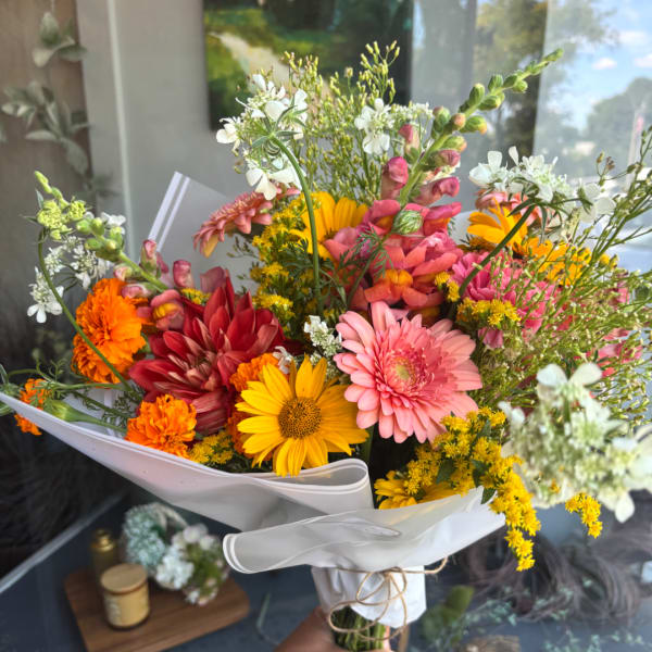 Handheld bouquet of mixed pink, yellow, orange, and white flowers wrapped in white paper