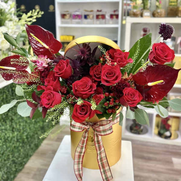 Red roses and dark red tropical blooms arranged in a gold hatbox with a plaid ribbon bow.