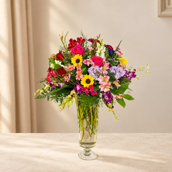 Mixed bouquet in a clear glass vase with bright pink, red, yellow, and purple flowers.
