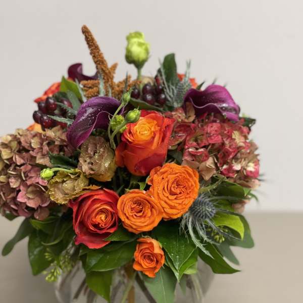 Low glass vase arrangement with bright orange and red roses, purple calla lilies, and antique hydrangeas