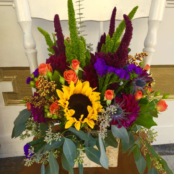 Colorful mixed bouquet with a sunflower, orange roses, and purple blooms in a rustic container.