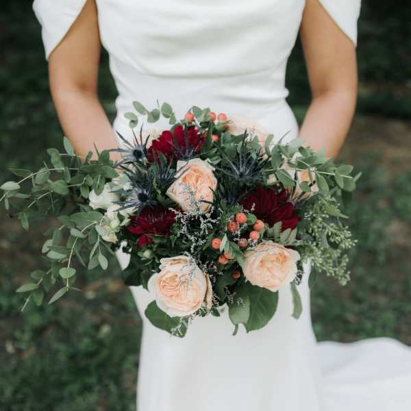 Bride holding a bouquet of peach and burgundy flowers with greenery