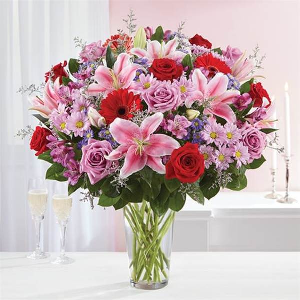 Mixed bouquet of pink lilies, red roses, and lavender flowers in a clear glass vase