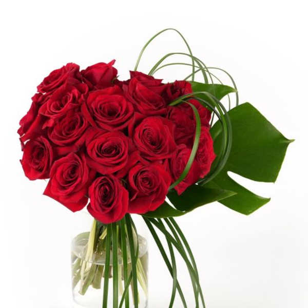 Tight bouquet of red roses with looping green blades in a clear glass cylinder vase