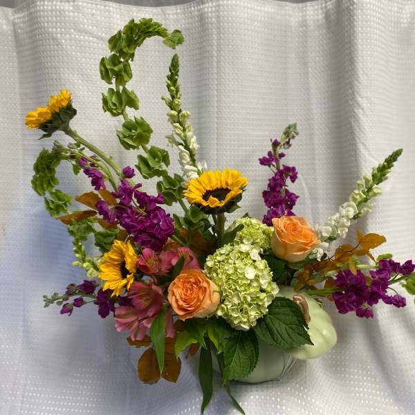 Mixed bouquet with sunflowers, peach roses, and purple blooms in a pale green pumpkin vase