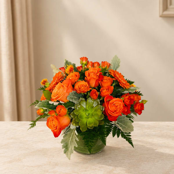 Orange roses and gerbera daisies in a glass vase with a green succulent accent.