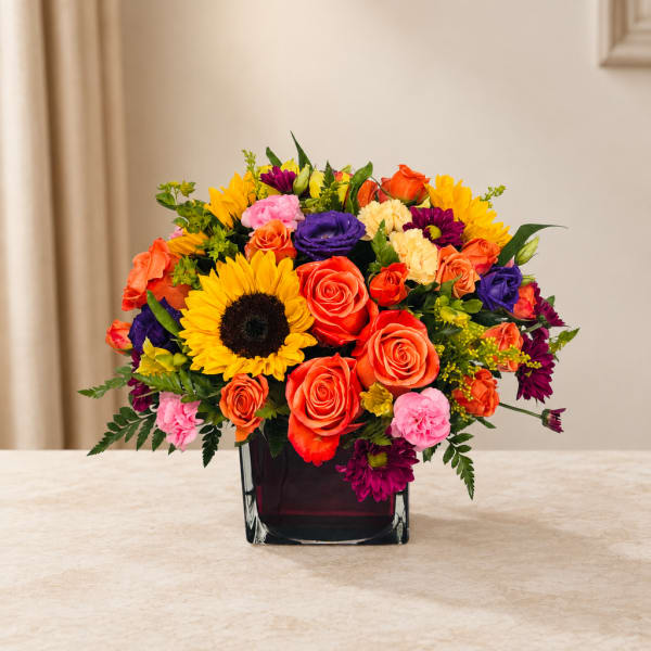 Colorful bouquet of roses, sunflowers, and carnations in a square glass vase