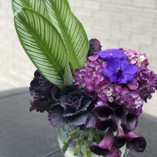 Purple hydrangea and dark calla lilies in a glass vase with striped green leaves
