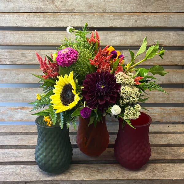 Three colorful flower arrangements in textured vases on a wooden bench